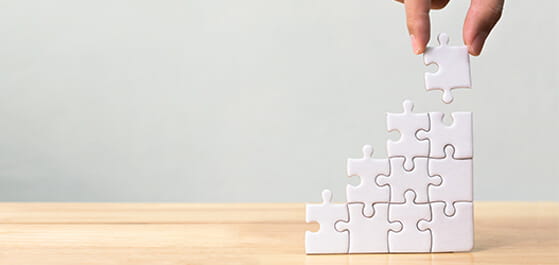 Header: Weiße Puzzleteile auf Holztisch stehend, von oben Finger halten  weiteres Puzzleteil.